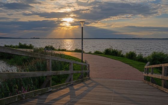 Eine Holzbrücke und anschliessender Weg führen durch einen See, im Hintergrund scheint die Sonne durch die Wolken am Himmel. 