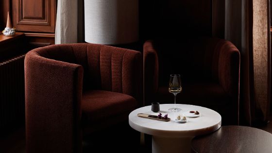 Zwei elegante, burgunderrote Samtsessel in einer Fensternische mit Vorhängen. Davor steht ein runder Beistelltisch, der mit verschiedenen kleinen Speisen und einem Glas Weißwein gedeckt ist.