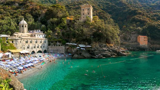 Belebter Sandstrand mit Sonnenschirmen vor einer mittelalterlichen Klosteranlage in einer kleinen Badebucht an einer mediterranen, bewaldeten Felsküste.