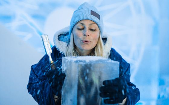 Eine Frau mit Mütze und dicker Jacke hält ein Schnitzmesser in der Hand, vor ihr steht ein grosser Eisblock. 