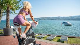 Rückansicht einer durchtrainierten Frau in lilafarbener Sportkleidung auf einem Spinning-Rad auf einer Terrasse an einem See bei Sonnenschein.