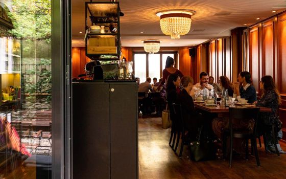 Personen sitzen in einem Restaurant an Tischen in einem holzverkleideten Gastraum mit Deckenleuchten aus Kristall, in einer Glastür spiegelt sich ein Außenbereich mit Sitzgelegenheiten im Garten.