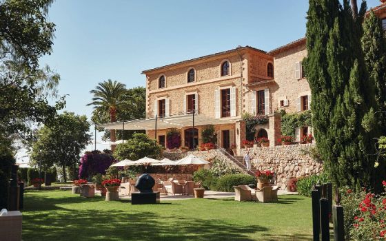 Ein sandfarbenes Gebäude mit Fensterläden uns Terrasse ist umgeben von Rasen, Palmen und grünen Pflanzen.