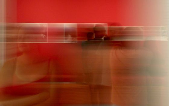 Langzeitbelichtung in einer Kunstgalerie mit roter Wand, unscharfen Personen und verschwommenen Bildern im Hintergrund.
