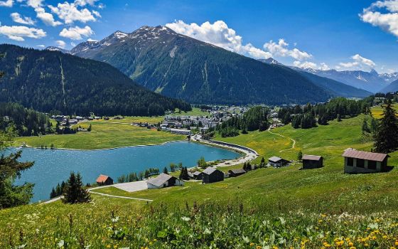 Mitten in einer Berglandschaft liegt ein kleiner See mit kleinen Häusern, im Hintergrund ragen Berge empor. 