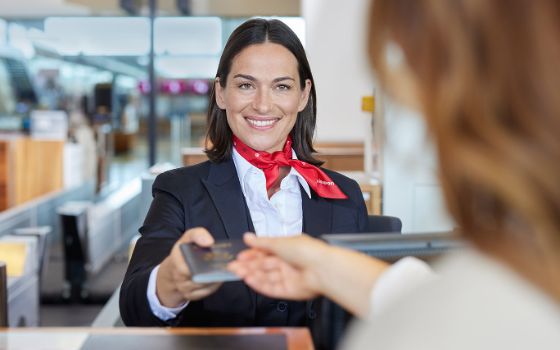 Porträt einer lächelnden Bodenstewardess mit einem roten Halstuch mit Swissport Logo, die den Reisepass einer Frau am Check-in-Schalter entgegennimmt.