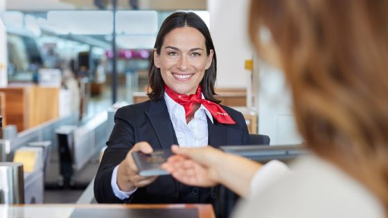 Porträt einer lächelnden Bodenstewardess mit einem roten Halstuch mit Swissport Logo, die den Reisepass einer Frau am Check-in-Schalter entgegennimmt.