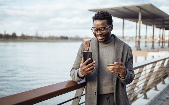 Ein Mann im grauen Mantel steht auf einem Steg an einem Geländer an einem Flussufer und schaut lächelnd auf ein Smartphone in seiner Hand, während er in der anderen eine Kreditkarte hält. 