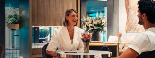 Eine Frau und ein Mann sitzen in einer eleganten Hotel-Lobby an einem Tisch, vor ihnen steht ein schicker Rollkoffer.