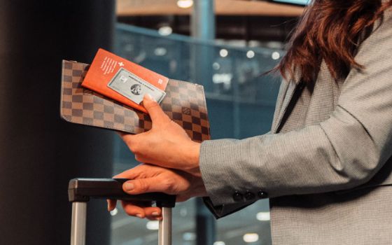 Eine silberne Platinum Kreditkarte von American Express mit Schweizer Reisepass in der Hand einer Frau mit Trolley in einem Flughafenterminal.