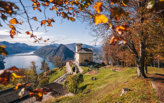 Herbstlicher Ausblick vom Monte Brè auf den Lago di Lugano mit Villa und farbigem Laub im Vordergrund.