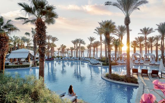 Luxuriöse, geschwungene Poollandschaft mit Palmen und zahlreichen Sonnenliegen vor einem Meerespanorama bei Sonnenuntergang. Im Vordergrund sitzt eine Frau in der Rückansicht auf einer der Liegen. 