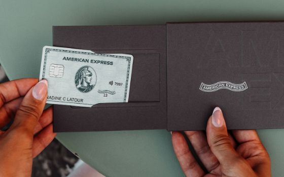 Sicht auf eine silberne Kreditkarte von American Express, die von einer Hand aus einer Box aus grauer Kartonage mit dem Aufdruck American Express auf einem Tisch gezogen wird