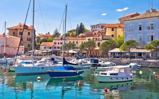 Verschiedene Boote liegen im Hafen eines mediterranen Ortes mit bunten Häusern an einer Uferpromenade bei Sonnenschein. 
