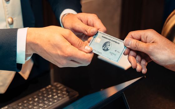 Nahaufnahme einer Hand, die eine platinfarbene Karte von American Express an eine Person in Uniform reicht, darunter verschwommen eine Tastatur. 
