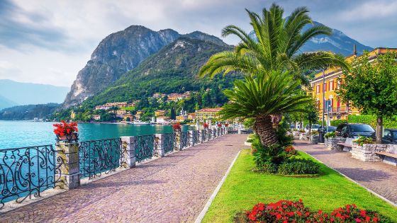 Promenade mit Palmen und roten Blüten an einem Seeufer in einer mediterranen Ortschaft vor Bergpanorama. 