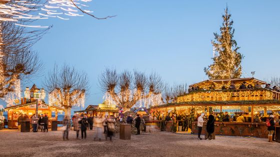 Personen auf einem Weihnachtsmarkt mit beleuchteten Holzhütten unter blauem Himmel am Abend an einem See. 