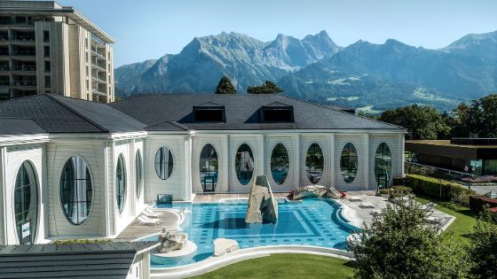 Neben einem weissen Gebäude mit ovalen Fenstern steht eine Außenpool mit Marmorskultur, im Hintergrund sieht man eine Berglandschaft. 