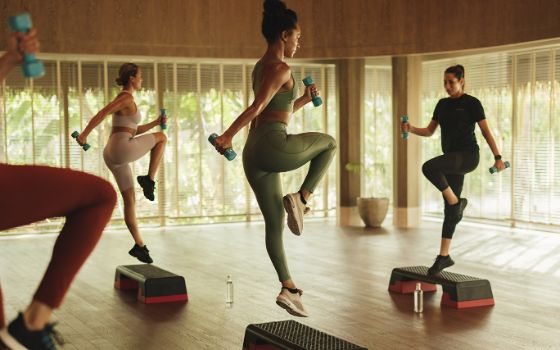 Drei Frauen machen mit einer Trainerin ein Workout mit Stepp und Kurzhanteln in einem Ressort. 
