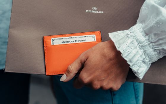 Eine Frau mit mit Steinen bestückten goldenen Armbaendern zahlt mit einer silbernen American Express Kreditkarte an einem Kartenlesegerät in der Hand einer Verkäuferin auf einem Sofa in einem Schmuckgeschäft.
