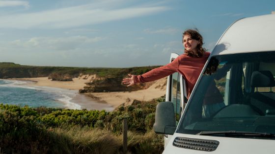 Eine lächelnde Frau steht mit ausgestrecktem Arm in der geöffneten Autotür eines Campervans auf einem Plateau an einer Meeresbucht mit Sandstrand. 