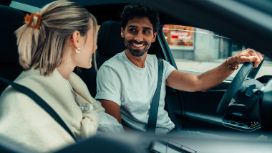 Ein Paar sitzt nebeneinander in einem Auto in urbaner Umgebung und schaut sich lächelnd an, er hat die Hand am Lenkrad.