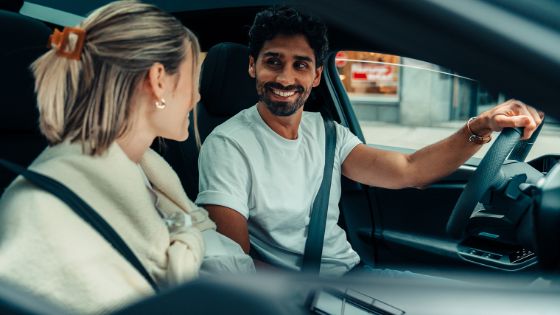 Ein Paar sitzt nebeneinander in einem Auto in urbaner Umgebung und schaut sich lächelnd an, er hat die Hand am Lenkrad.