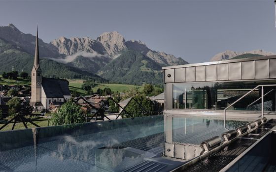 Moderner Gebäudekomplex mit Outdoor-Pool vor einer Ortschaft mit Kirche, umgeben von grünen Wiesen vor Bergpanorama. 
