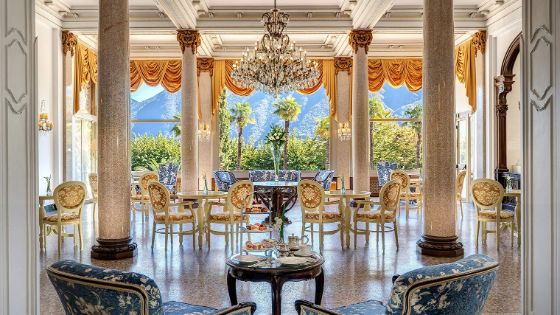 In einer Hotelhalle stehen verzierte Säulen, Stühle mit Blumenmuster sind um Tische platziert und an der Decke hängt ein grosser Kronleuchter. Im hinteren Bereich ist eine grosse Fensterfront, dahinter eine Berglandschaft.