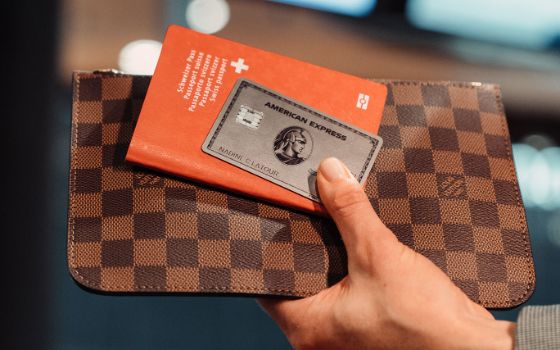 Nahaufnahme einer silbernen Kreditkarte von American Express auf einem roten Reisepass vor einem braunem Lederetui, zusammen hochgehalten von einer Hand 