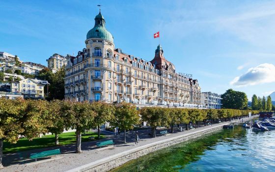 Blick auf das Hotel Mandarin Oriental in Luzern, davor Wasser und ein Weg, der von Bänken und Bäumen gesäumt ist. 