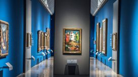 An einer Säule hägt ein Gemälde in einem Goldrahmen, die Säule steht in einem langen Gang mit blauen Wänden, an denen weitere Gemälde hängen.