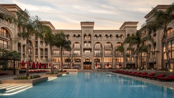 Luxuriöse Poollandschaft mit roten Sonnenliegen und Palmen im Innenhof eines palastartigen Hotelkomplexes mit orientalischen Bauelementen. 