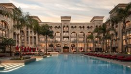 Luxuriöse Poollandschaft mit roten Sonnenliegen und Palmen im Innenhof eines palastartigen Hotelkomplexes mit orientalischen Bauelementen. 