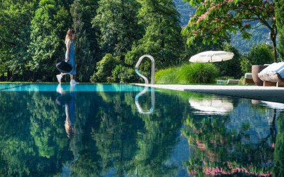 Eine Frau in sommerlichem Kleid geht am Rande eines Schwimmbeckens entlang. Hinter dem Pool befinden sich grüne Bäume und Büsche, ein Sonnenschirm und eine Liege.  