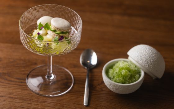 In einem Stielglas ist ein Macaron mit einer Mousse dekoriert, daneben steht eine weisse Kugel mit grünen Körnern.
