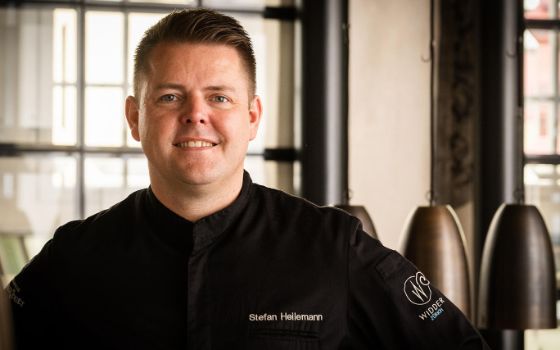 Porträt eines Mannes in schwarzer Kochjacke mit der Aufschrift «Stefan Heilemann» und einem Logo in einem Raum. 