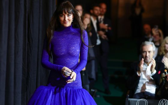 Dakota Johnson geht in einem eleganten blauen Kleid auf eine Bühne während hinter ihr das Publikum applaudiert. 
