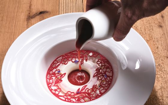 Eine Hand lässt einen Klecks roter Sauce aus einer kleinen Karaffe auf einen Teller tropfen, in dem eine weisse Creme mit Dekoration ist. 