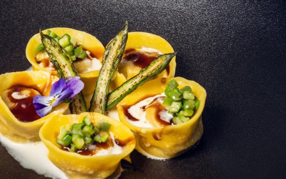 Auf einem schwarzen Teller liegen 5 Ravioli, die mit brauner und weisser Sosse beträufelt sind. In der Mitte sind drei Spargelspitzen und ein violettes Veilchen als Dekoration. 