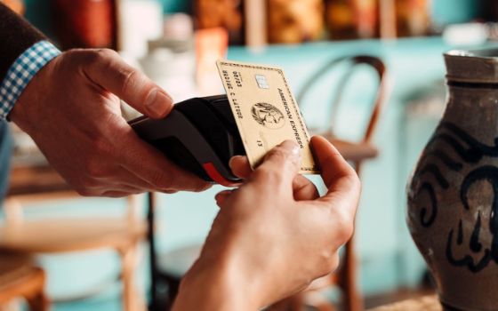 Nahaufnahme einer Hand, die eine goldene Kreditkarte von American Express an ein Kartenlesegerät hält, dieses wird von einer zweiten Person gereicht. Daneben eine Vase, im Hintergrund sind verschwommen Stühle zu sehen.
