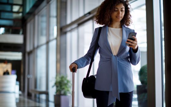 Eine junge Frau steht im Flughafengebäude und schaut auf ihr Smartphone, sie hält den Griff ihres Reisekoffers. 