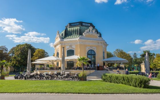 Gelber Pavillon mit opulenter Stuck-Verzierung an der Aussenfassade bei Sonnenschein und blauem Himmel, davor aufgespannte Sonnenschirme und Tischgruppen mit Stühlen. 