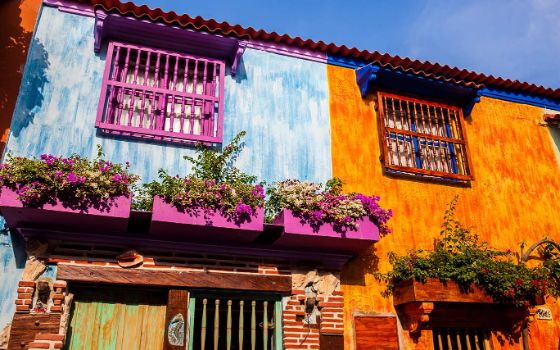 Ein blaues und ein orangenes Haus stehen direkt nebeneinander, beide haben verzierte, bunte und verschnörkelte Gitter vor den Fenstern und Türen, darüber hängen üppigen Blumenkästen. 