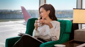Eine stilvoll gekleidete Frau sitzt mit einer Tasse Kaffee in den Händen in einem grünen Sessel an einer Glasfassade in einer Flughafen Lounge und blickt entspannt hinaus aufs Rollfeld unter blauem Himmel.