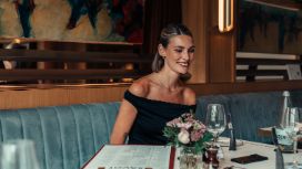 Eine Frau trägt ein schwarzes, schulterfreies Kleid und sitzt hinter einem gedeckten Tisch auf einem hellblauen Sofa.