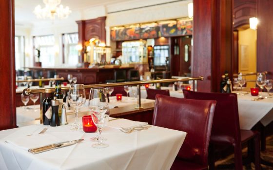 Speisesaal eines edlen Restaurants mit weiss eingedeckten Tischen und weinroten Einrichtungselementen, im Hintergrund ein Barbereich.