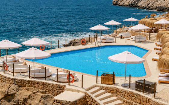 In einer Meeresbucht steht ein Swimmingpool mit Liegen und Sonnenschirmen. 