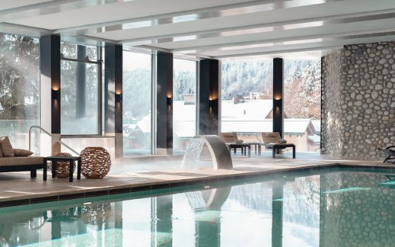 Elegante Polsterliegen in einem Spa-Bereich mit Pool und Panoramafenstern mit Blick auf eine verschneite Ortschaft in den Bergen.