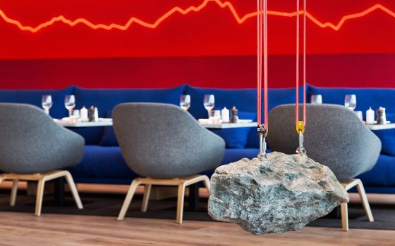 Vor einer roten Wand stehen blaue Sofas mit Tischen und grauen Sesseln, von der Decke hängt ein grosser Stein an Karabinerharken und Seilen.